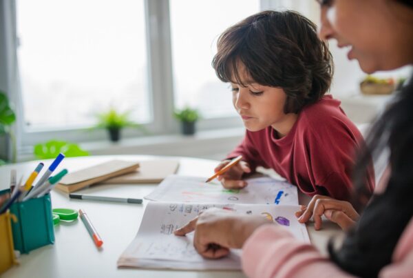 A child sits at a desk doing homework with the help of an adult, who is pointing at a page in a workbook. Colored pencils and school supplies are scattered on the table near a bright window.