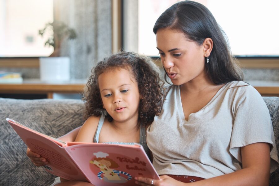 A woman and a young girl sit closely on a couch, reading a colorful children’s book together. Both are engaged with the story, appearing relaxed and focused.