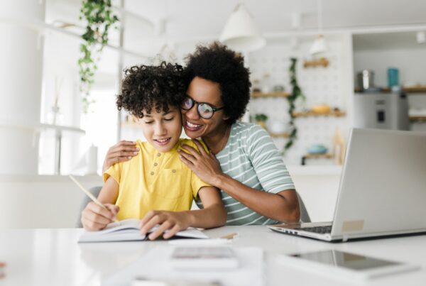 A smiling woman hugs a child who is writing in a notebook at a table with a laptop in a bright, modern kitchen. Both appear happy and engaged in the activity.