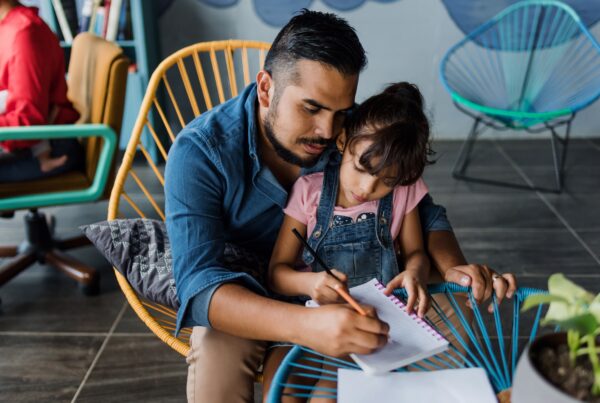 A man sits closely with a young girl on his lap, helping her write in a notebook at a round table. Both appear focused, and the setting is indoors with colorful chairs in the background.