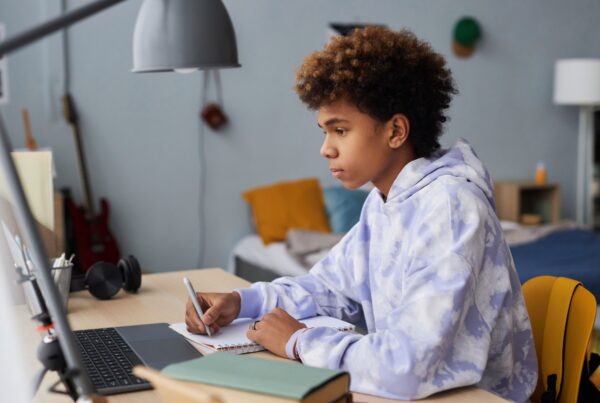 A teenage student wearing a tie-dye hoodie sits at a desk, writing in a notebook while looking at a laptop screen in a bedroom. The room has a bed, a guitar, and books visible in the background.