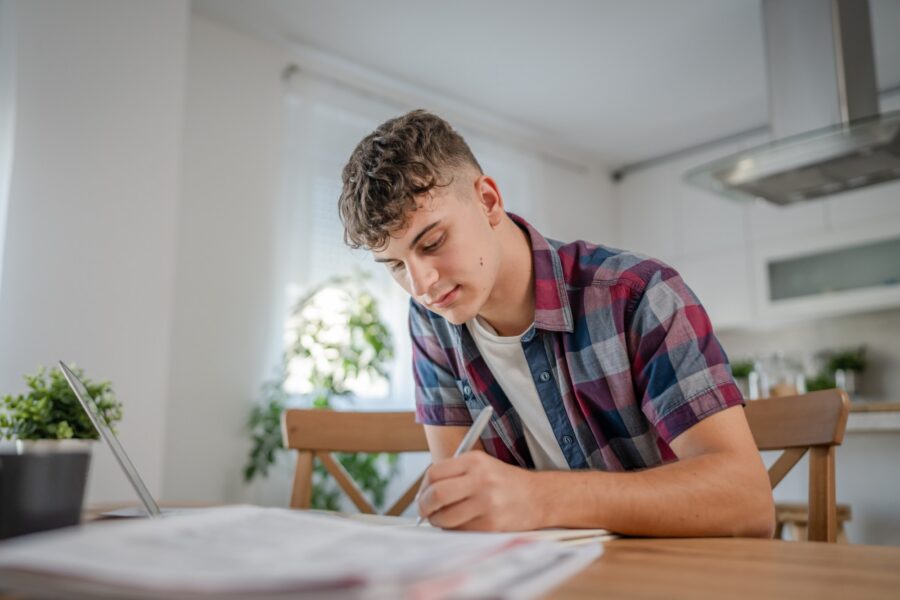 A young man with curly hair sits at a table in a bright kitchen, writing in a notebook with a pen, while a laptop and papers are spread out in front of him.