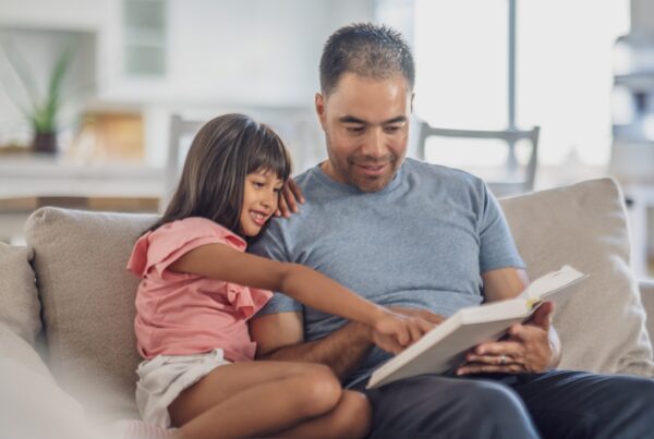 A young girl and an adult man sit together on a couch, smiling and reading a book. The girl is pointing at something on the page while the man holds the book and looks on, both appearing happy and engaged.