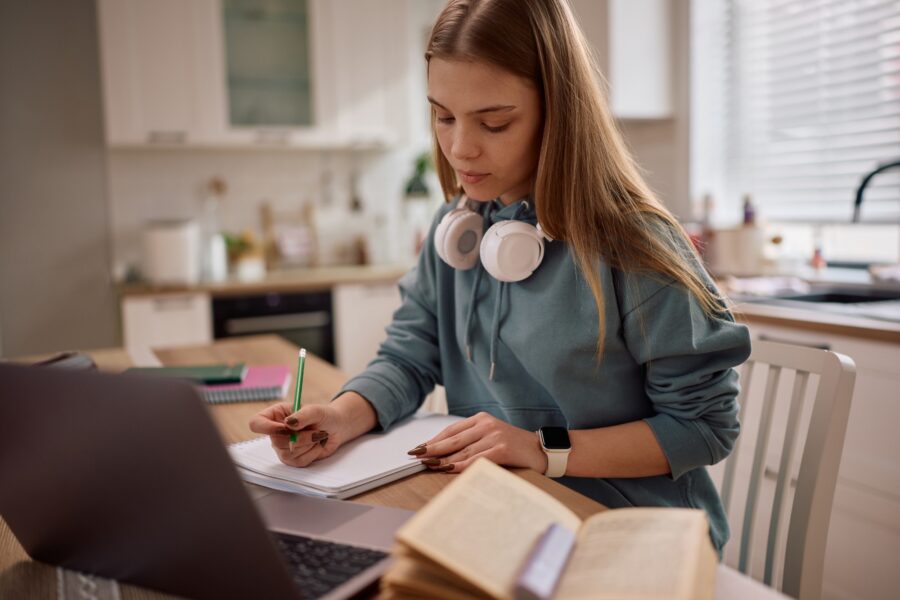 A young woman with headphones around her neck writes in a notebook while studying at a kitchen table with an open laptop and book in front of her. She appears focused and is surrounded by study materials.