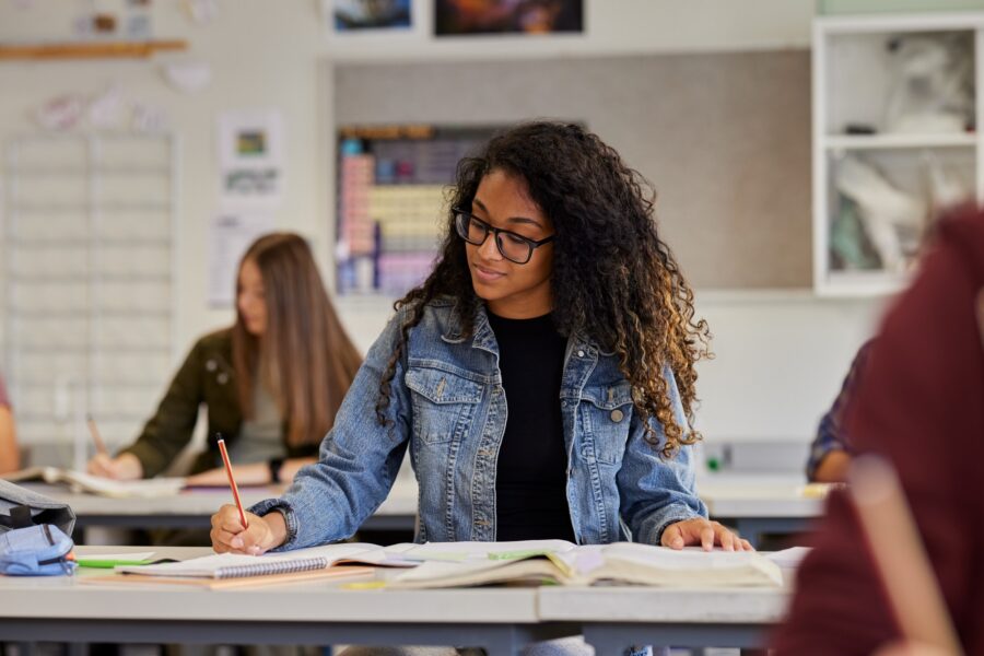 A student with curly hair and glasses, wearing a denim jacket, sits at a desk in a classroom writing in a notebook with open books in front of her. Other students are also seated and working in the background.