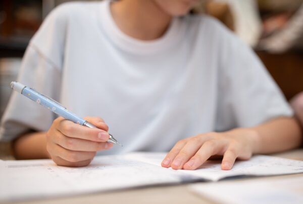 A person wearing a white shirt is writing with a blue pen while studying or doing homework, with an open notebook or workbook in front of them. The focus is on their hands and the pen.