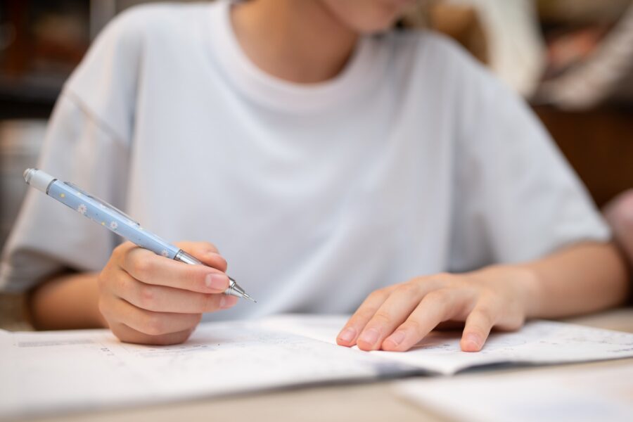 A person wearing a white shirt is writing with a blue pen while studying or doing homework, with an open notebook or workbook in front of them. The focus is on their hands and the pen.