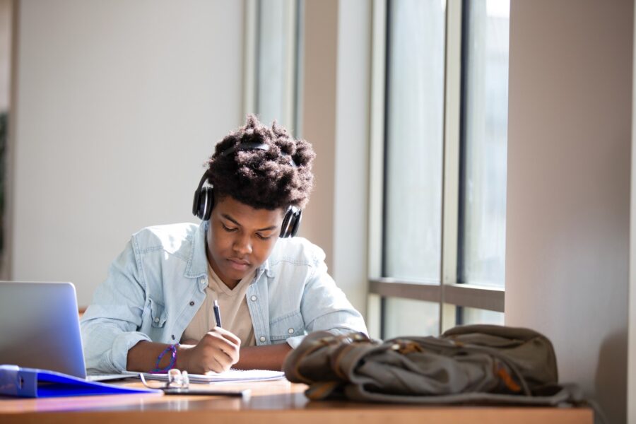 A student wearing headphones sits at a desk by a window, writing in a notebook. A laptop, a blue folder, and a backpack are on the desk beside him. Sunlight streams in through the large windows.