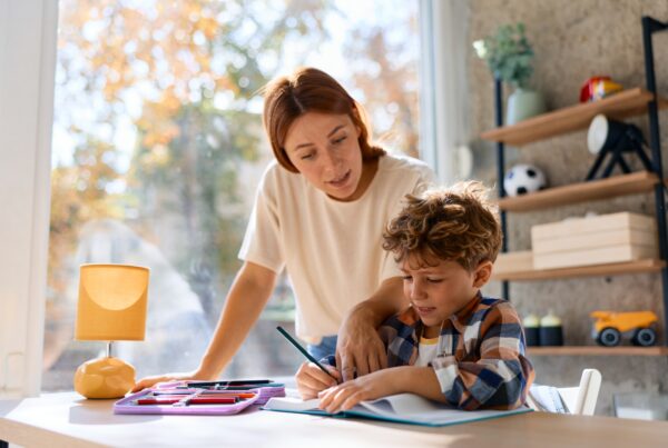 A woman helps a young boy with his homework at a desk by a window. Art supplies and a lamp are on the desk, and shelves with toys are seen in the background. Sunlight streams into the room.