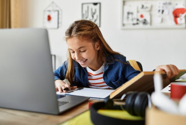A smiling young girl sits at a desk using a laptop, writing in a notebook. Art supplies, books, and headphones are on the desk. Childrens drawings hang on the wall in the background.