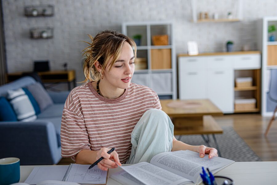 A young woman sits at a table in a living room, studying with open books and notes. She holds a pen and appears focused, with a cozy, modern background featuring shelves and a sofa.