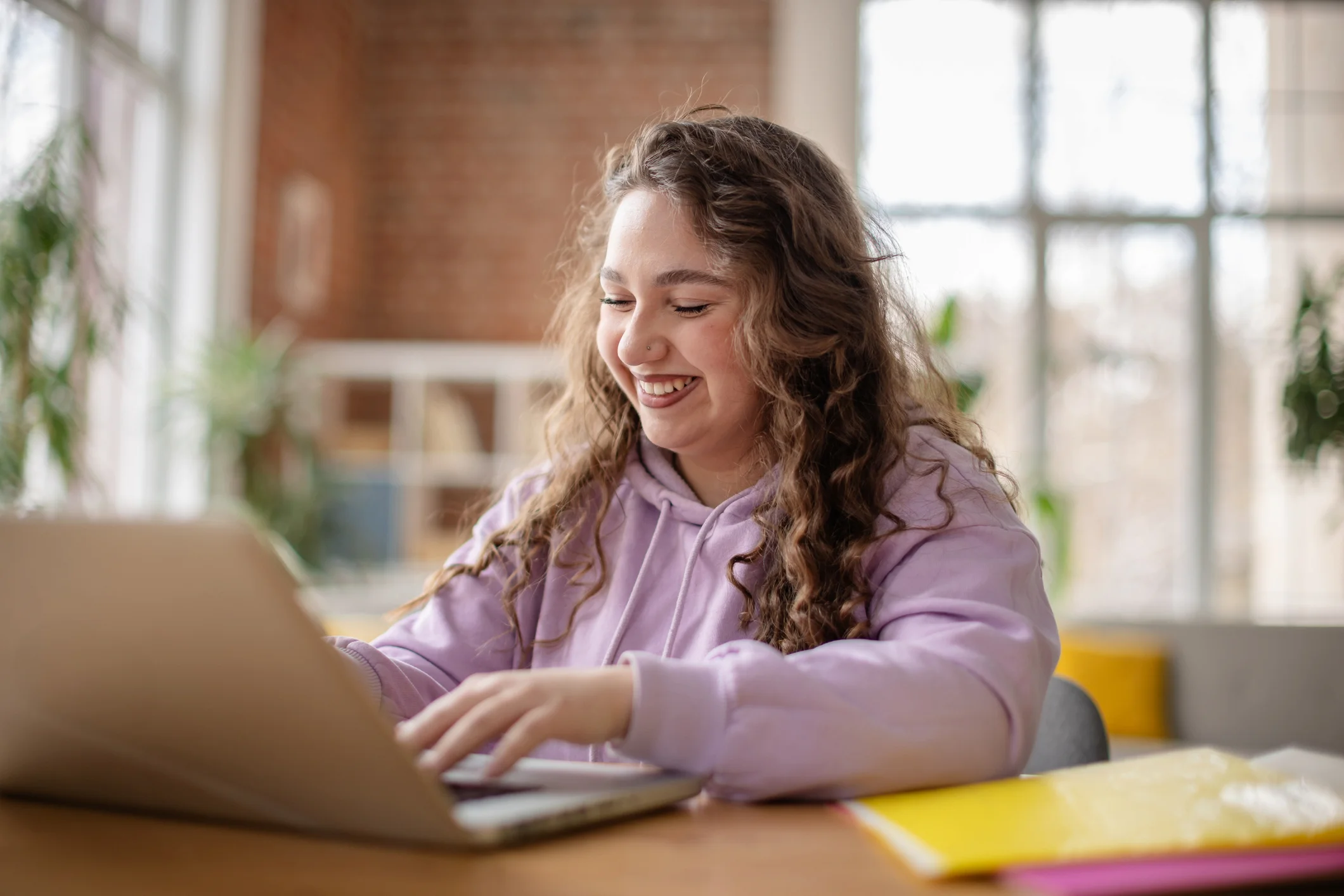 A smiling young woman with curly hair, wearing a lavender hoodie, sits at a table using her laptop for an Online Algebra Tutors session. She appears happy and engaged, with notebooks and plants in the bright background.